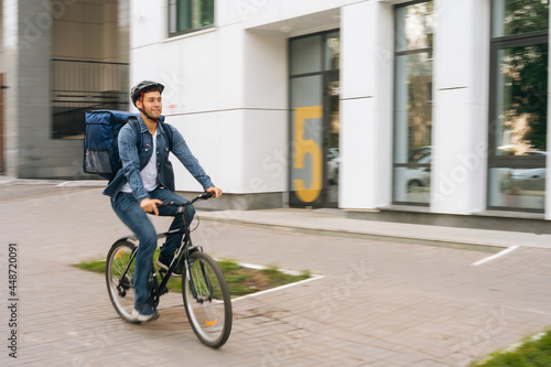 Cheerful handsome young delivery man with thermo backpack riding bicycle in city street on blurred background of office building. Courier male wearing protective helmet delivery food to client