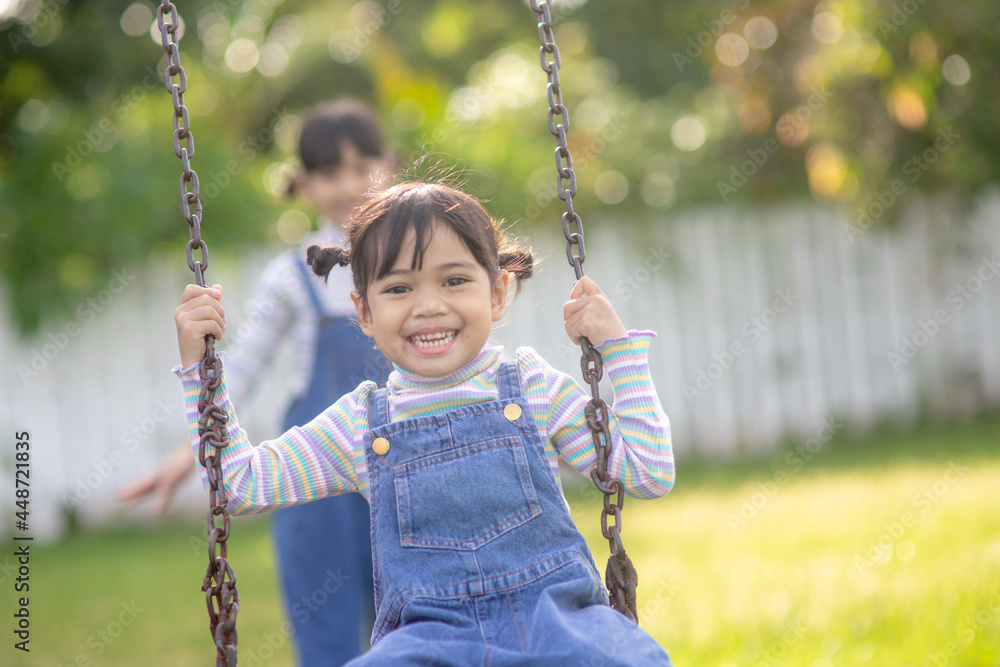 Two cute little sisters having fun on a swing together in a beautiful summer garden on a warm and sunny day outdoors. Active summer leisure for kids. Two cute little sisters having fun on a swing together in a beautiful summer garden on a warm and sunny day outdoors. Active summer leisure for kids.