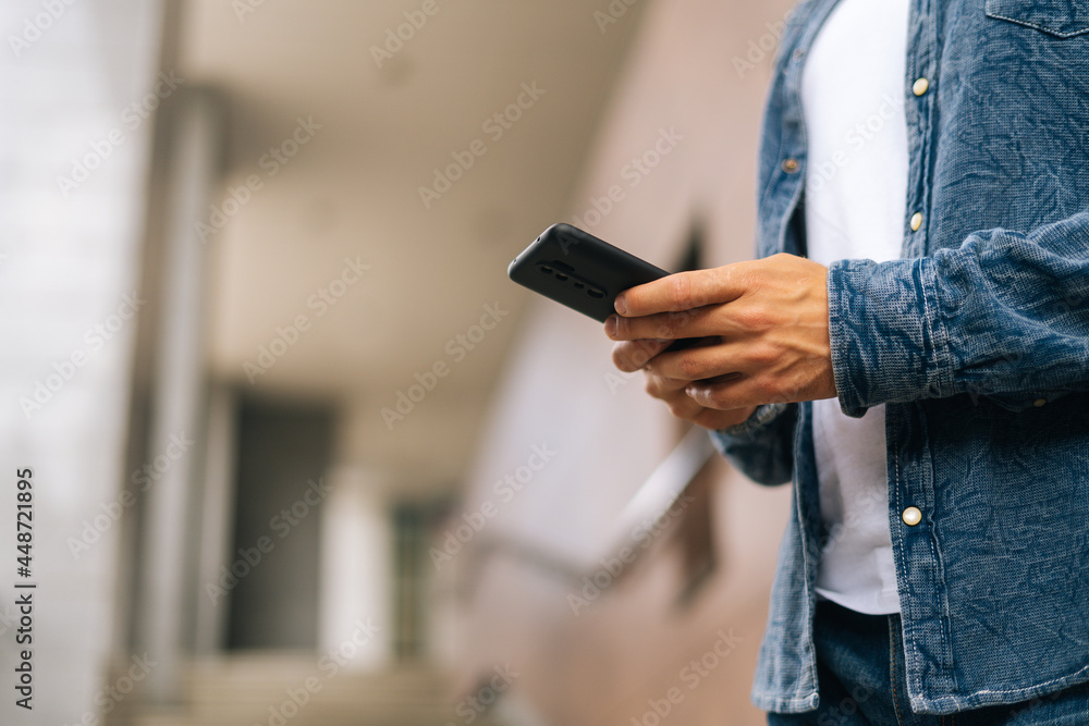 Close-up hands of unrecognizable man texting message on mobile phone outdoors on blurred background, side view, selective focus. Closeup view of male typing sms on modern smartphone outdoors.