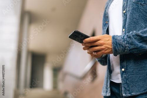Close-up hands of unrecognizable man texting message on mobile phone outdoors on blurred background  side view  selective focus. Closeup view of male typing sms on modern smartphone outdoors.