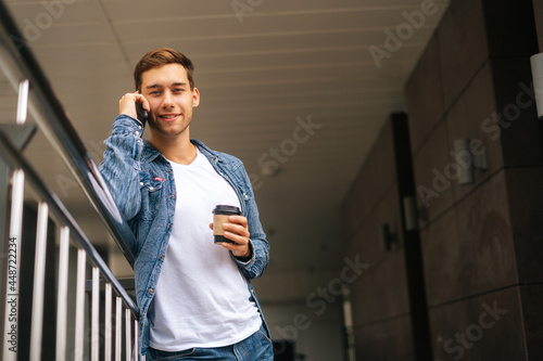 Low-angle view of smiling young man talking on mobile phone, standing on balcony terrace with coffee coffee cup leaning on railing of office building, blurred background, looking at camera.