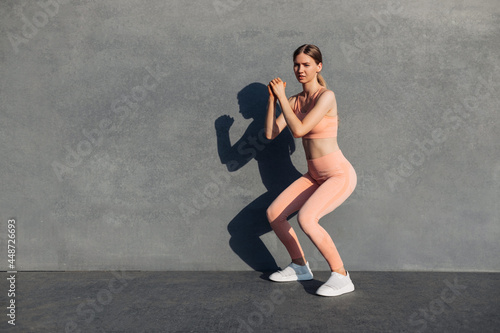 Side view shot of fit young woman doing squats, exercise on buttocks outdoors, workout on urban concrete wall background