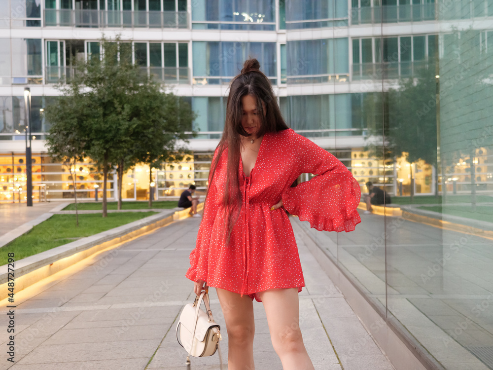 Fototapeta premium Attractive smiling Asian girl in red dress poses next to glass facades of buildings.
