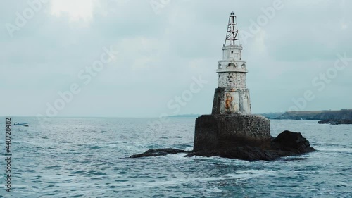 Sea waves hitting and splashing against old white lighthouse and dark gray cliffs. Isolated on blue cloudy sky. Fishing boat passing. Near town of Ahtopol on Black sea coast, Bulgaria