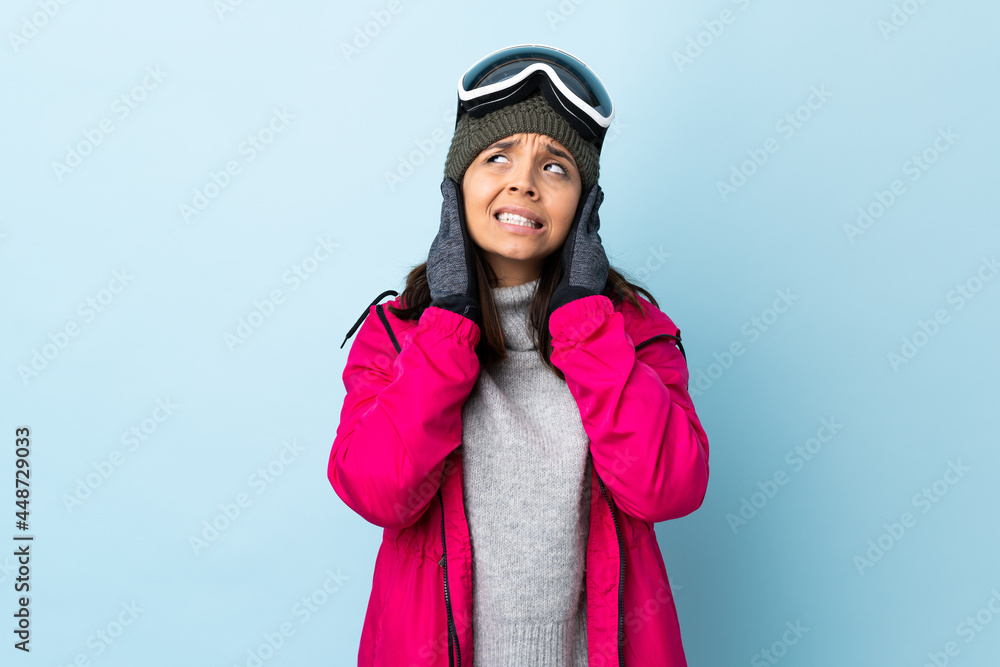 Mixed race skier girl with snowboarding glasses over isolated blue background frustrated and covering ears. Mixed race skier girl with snowboarding glasses over isolated blue background frustrated and covering ears.