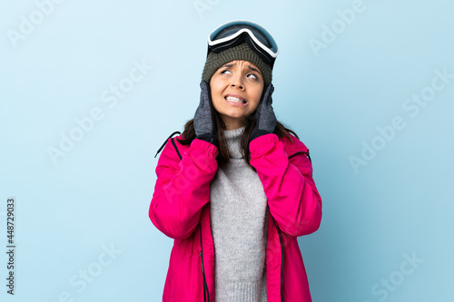 Mixed race skier girl with snowboarding glasses over isolated blue background frustrated and covering ears.