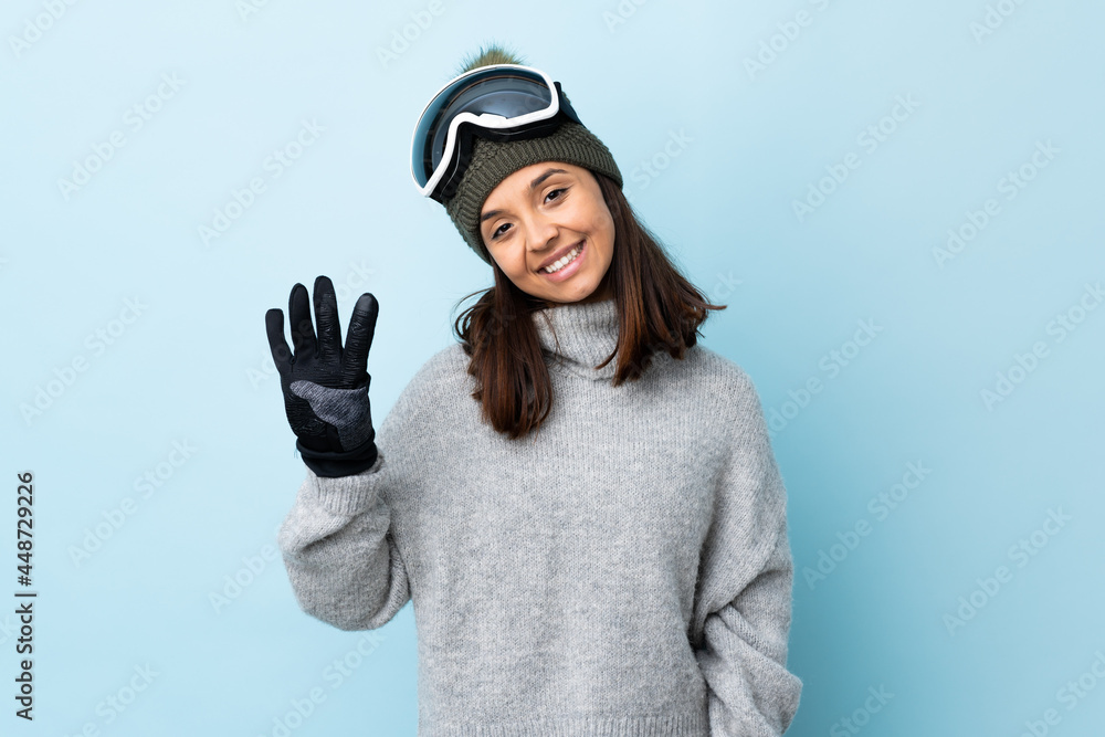 Mixed race skier girl with snowboarding glasses over isolated blue background happy and counting four with fingers. Mixed race skier girl with snowboarding glasses over isolated blue background happy and counting four with fingers.