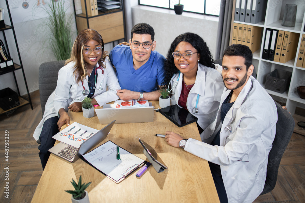 Top angle view of multiracial group of doctors wearing coats and scrubs sitting together at table and smiling on camera. Medical specialists during session at hospital. Cooperation concept.
