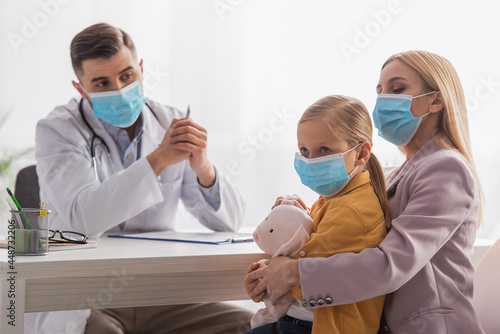Kid looking away near mother in medical mask and family doctor in clinic