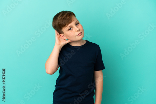 Little redhead boy isolated on blue background listening to something by putting hand on the ear