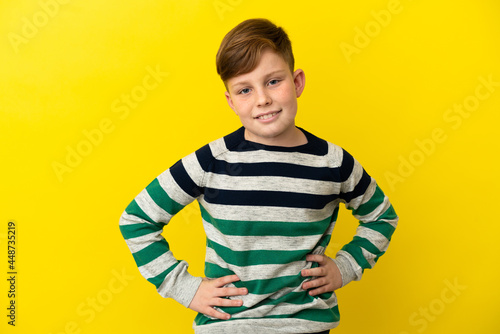 Little redhead boy isolated on yellow background posing with arms at hip and smiling