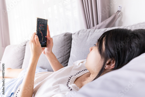 Young Asian woman lying on sofa and looking at financial stock market charts rate dynamic, trading data indexes growth using mobile phone for cryptocurrency digital analytics.
