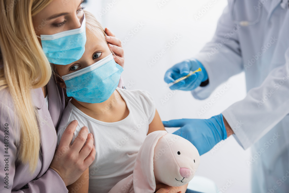 Mother hugging kid in medical mask near pediatrician with syringe on blurred background Mother hugging kid in medical mask near pediatrician with syringe on blurred background