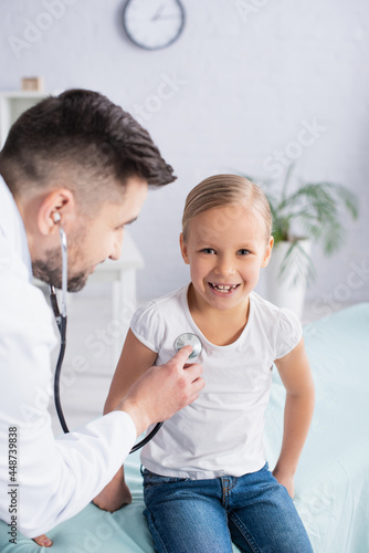 Blurred pediatrician using stethoscope on cheerful kid in clinic