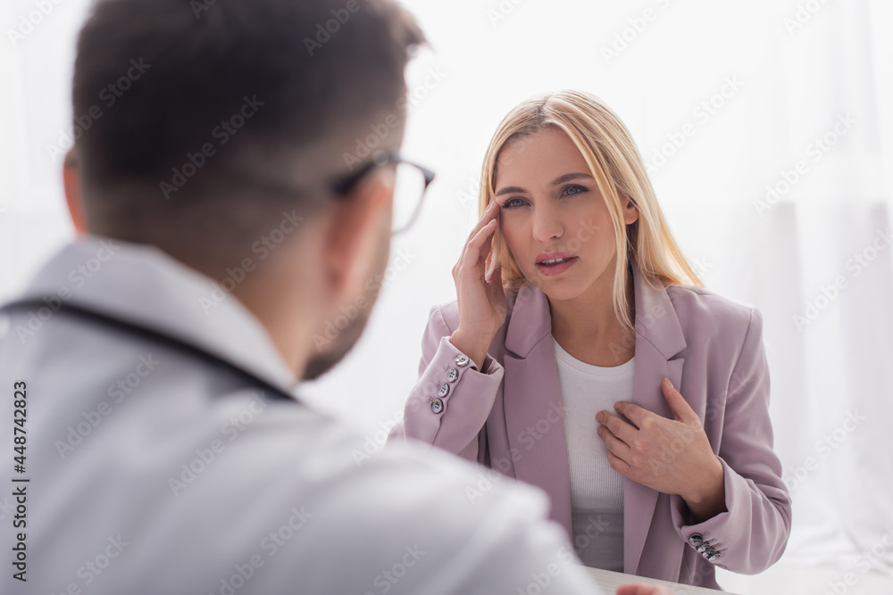 woman touching head and chest while talking to blurred doctor during consultation woman touching head and chest while talking to blurred doctor during consultation