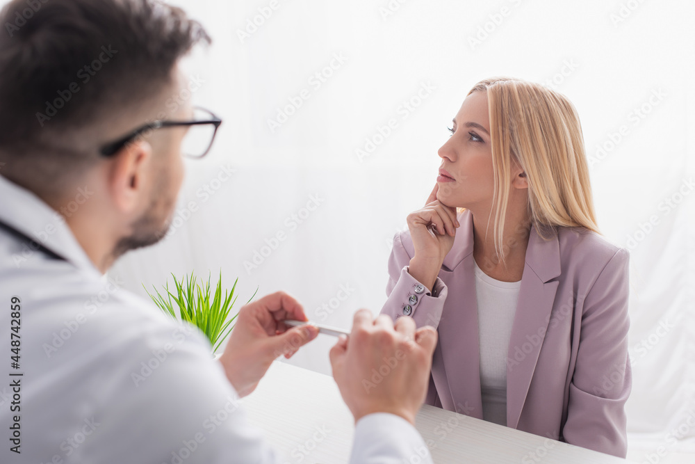 blonde woman thinking near blurred doctor during appointment in clinic