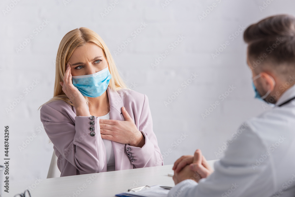 sick woman in medical mask touching head and chest during consultation with blurred doctor