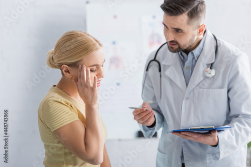 doctor with clipboard talking to woman touching head with closed eyes