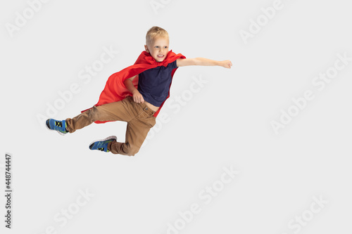 Portrait of little blond boy, pupil flying like super hero in red cape isolated on white studio background. Copyspace for ad. Childhood, education, emotion concept.