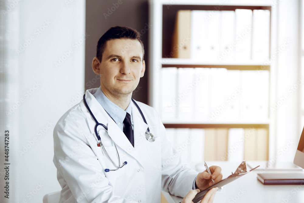 Friendly male doctor sitting and working with clipboard of medication history records in sunny clinic. Medicine and healthcare concept Friendly male doctor sitting and working with clipboard of medication history records in sunny clinic. Medicine and healthcare concept