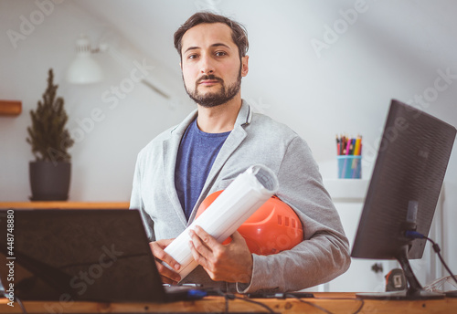 A man with drawings and a helmet poses for a photo while sitting at a table with a laptop and an additional monitor