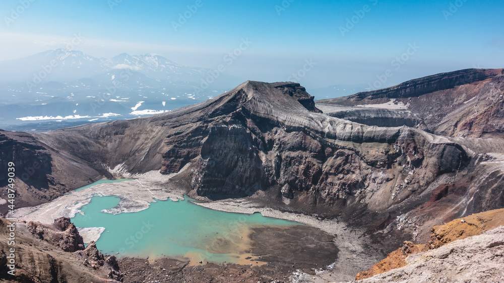 An unusual acid lake on the top of the volcano is surrounded by steep rocky slopes. Lifeless turquoise water with sulfur deposits. A snow-covered mountain range  in the haze. Kamchatka. Gorely Volcano