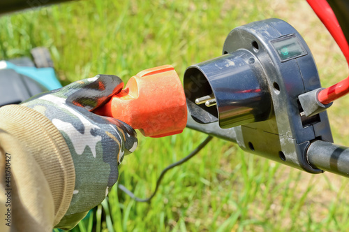 Gloved hand with electric cable and plug connects the lawn mower to the power supply. Electricity and people, safety when using electrical appliances in the garden and backyard.