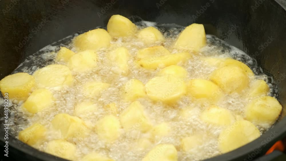 Potato is frying in cauldron in boiling hot oil on nature on fire, closeup view. Large pieces of potatoes are deep-fried in a black kazan. Cooking using wood-burning stove.