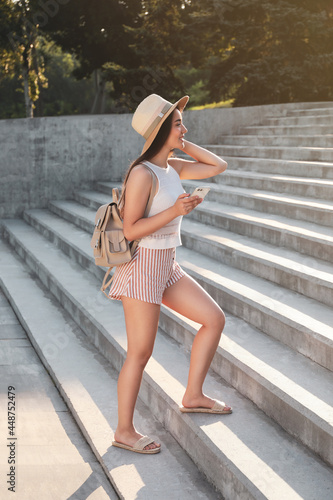 Beautiful young woman with stylish backpack and smartphone walking up stairs outdoors