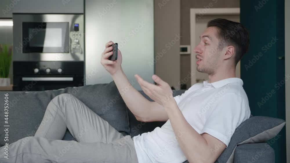 Smiling young man with smartphone making video call while lying on couch in living room. Portrait of happy man mobile video calling, resting on sofa at home Smiling young man with smartphone making video call while lying on couch in living room. Portrait of happy man mobile video calling, resting on sofa at home