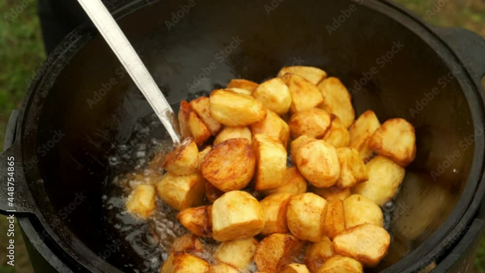 The man takes out of the cauldron ruddy tasty potato using spatula, closeup view. Potato is frying deep-fried in a black kazan. Cooking using wood-burning stove on nature. The man takes out of the cauldron ruddy tasty potato using spatula, closeup view. Potato is frying deep-fried in a black kazan. Cooking using wood-burning stove on nature.