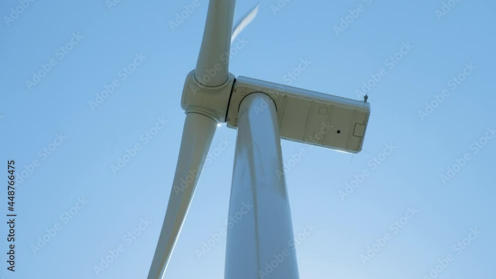 White wind driven generator with huge rotating blades generating electricity stands against clear blue sky close low angle shot White wind driven generator with huge rotating blades generating electricity stands against clear blue sky close low angle shot