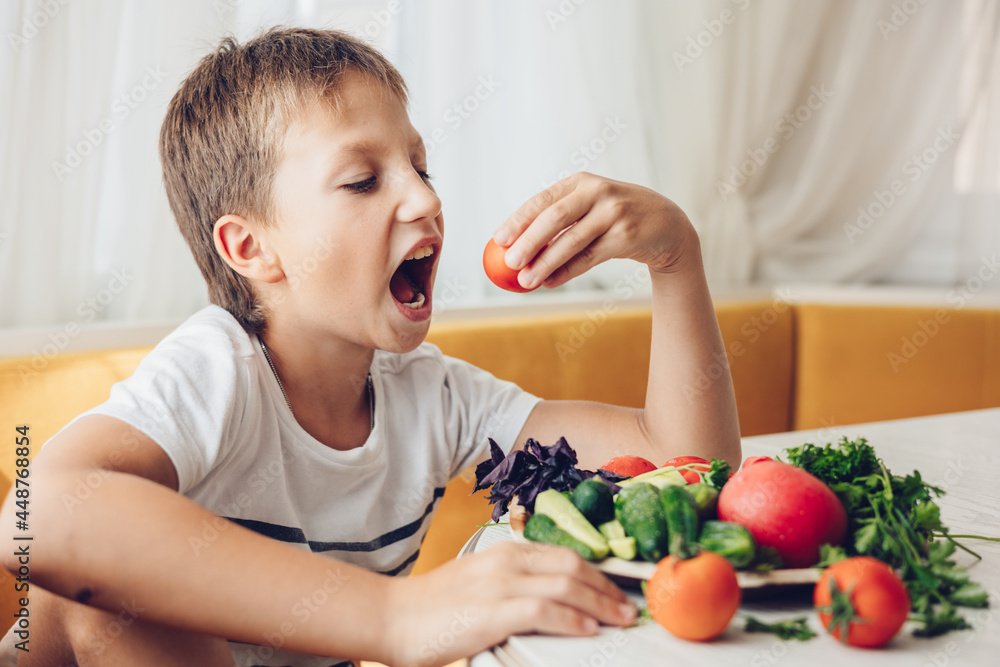 Boy sitting in the kitchen eating healthy vegetables cucumber and tomato Boy sitting in the kitchen eating healthy vegetables cucumber and tomato