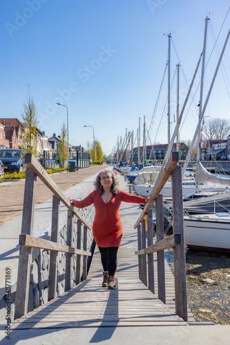 Mature woman standing on a wooden ramp in the port of Brouwershaven boats in the background, brown casual clothes and black tights with hiking boots in Schouwen-Duiveland in Zealand, Netherlands