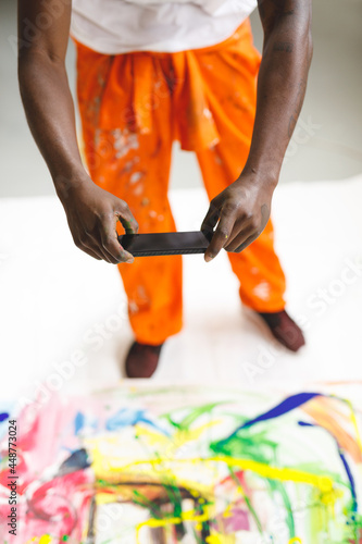 African american male painter at work taking picture of artwork with smartphone in art studio