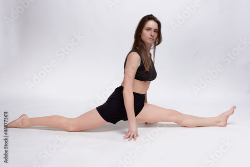 Young slim athletic girl, with long hair, in a black top and shorts, in the studio on a white background, does the splits