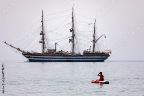 Italy. Scauri. A young man is engaged in SUP-surfing against the background of the sailboat Amerigo Vespucci.