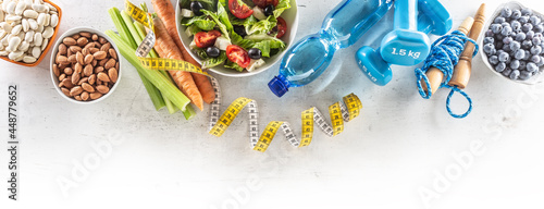 Healthy fresh salad with tomatoes surrounded with exercise equipment, carrtot celery and measuring tape - top of view