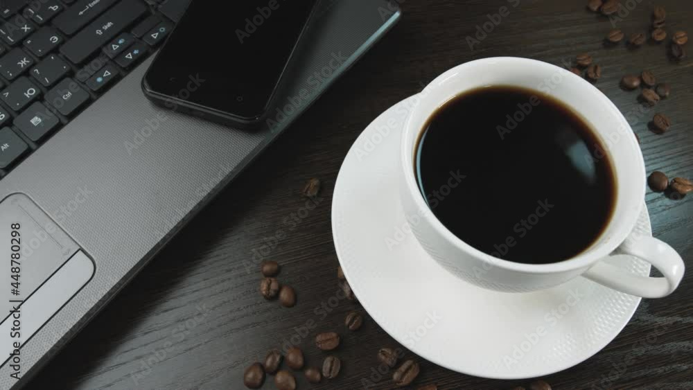 The camera moves smoothly over the table, on which there is a cup of coffee on a saucer. Next to it is an open laptop and a cell phone. Coffee beans are also scattered on the table. The camera moves smoothly over the table, on which there is a cup of coffee on a saucer. Next to it is an open laptop and a cell phone. Coffee beans are also scattered on the table.