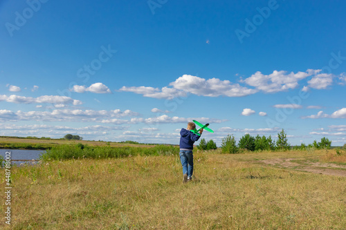 A six-year-old preschooler boy in a blue jacket launches a toy plane in a field against a blue sky with clouds on a summer day. The bright sun is shining. Scenery © ALEKSEI
