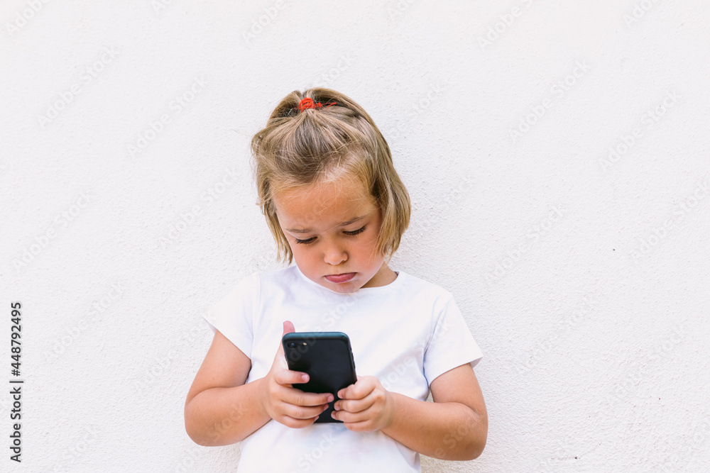 Little blond hair girl wearing white t-shirt, looking at mobile phone, with concentrated face Little blond hair girl wearing white t-shirt, looking at mobile phone, with concentrated face