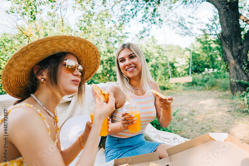 Two women sitting on a sofa in the backyard and eating pizza, drinking lemonade on summer sunny day. 