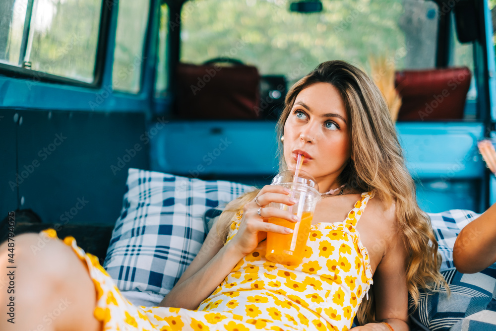 Portrait of young pretty woman sitting on blanket in a van and drinking lemonade outdoors in nature. Enjoying summer, travel concept - obrazy, fototapety, plakaty 