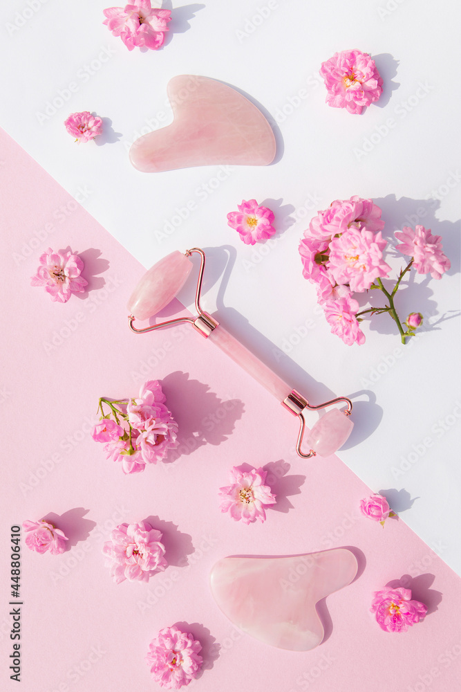 Facial massage kit for home spa. Face roller  and gua sha massager made from rose quartz on white and pink pastel background with rose buds. Natural treatment concept.