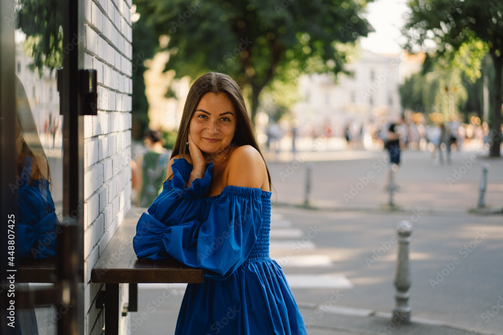 Fototapeta premium Portrait happy young woman wearing blue dress laughing looking at camera standing on street. Urban background