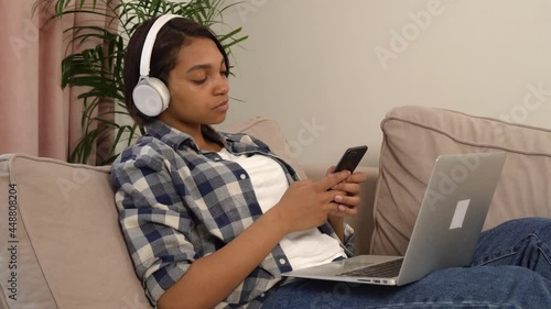 A young African-American woman lying on the couch with a laptop listening to music in headphones with a phone in her hands. The concept of a holiday home.