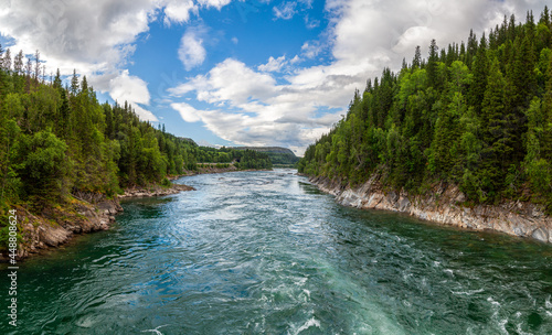 Frontal panorama of a wide river among the taiga forest on steep banks under blue skies in Norway in the summer.