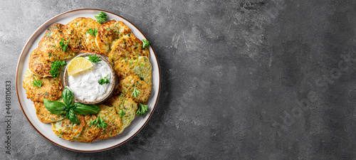 Zucchini pancakes with herbs and sour cream on a plate and gray background top view, web banner with copy space for text. Vegetarian dish