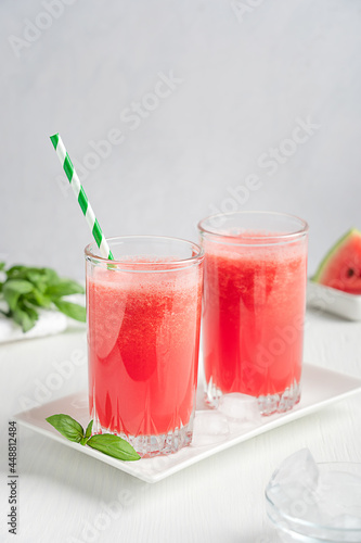 Watermelon refreshing juice served in two drinking glasses with paper straw and green basil leaves on plate on white wooden background with fruit slices as dessert at kitchen in summer. Vertical image