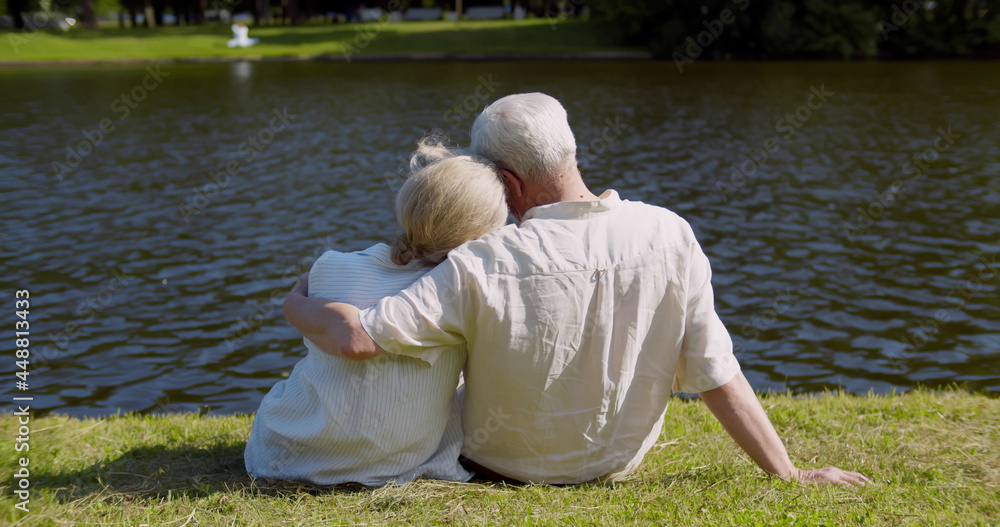 Rear view of senior couple sitting on grass near river and embracing Rear view of senior couple sitting on grass near river and embracing